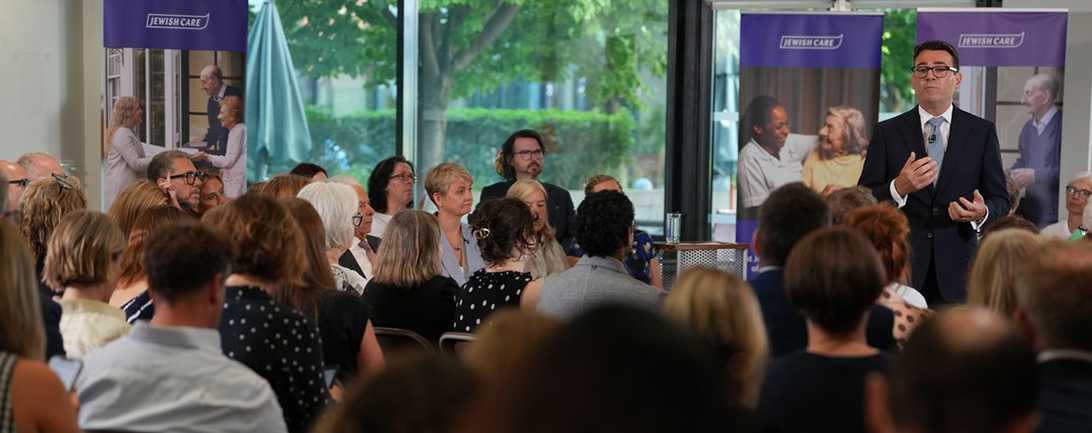 UK Prime Minister Andy Burnham speaking at a podium during a social care event, addressing a seated audience in front of promotional banners.