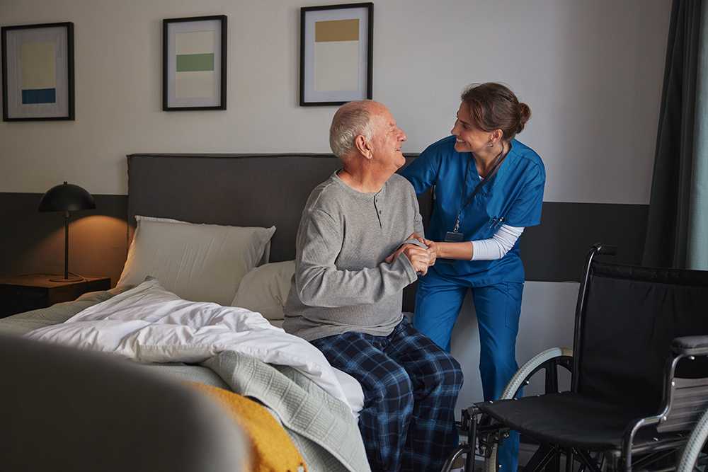 Carer in blue helping old man off bed in council run care home