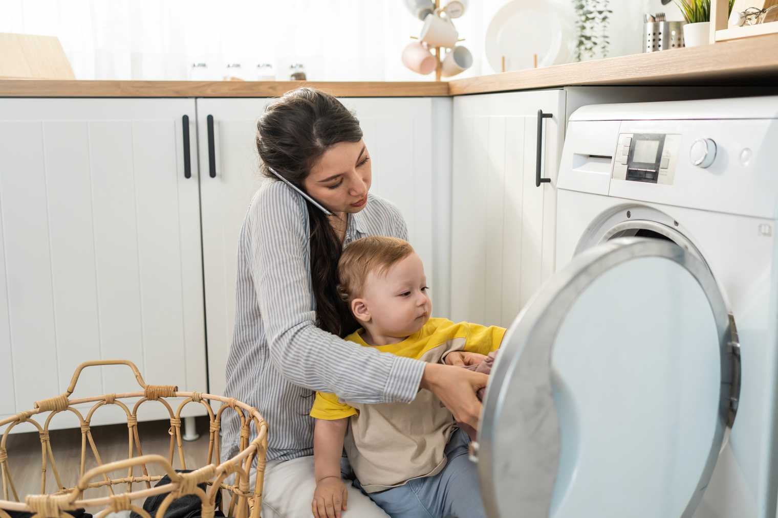 woman working on the phone while doing laundry and also looking after her infant son