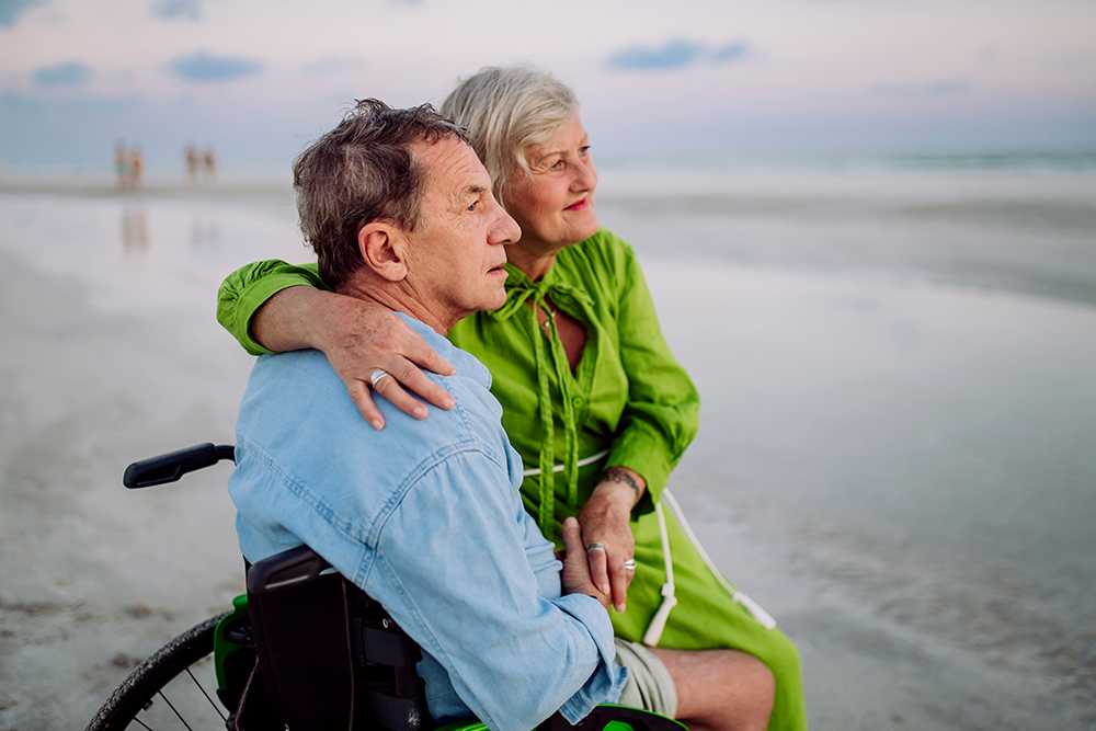 An elderly man in a wheelchair and old lady cuddling him on the beach