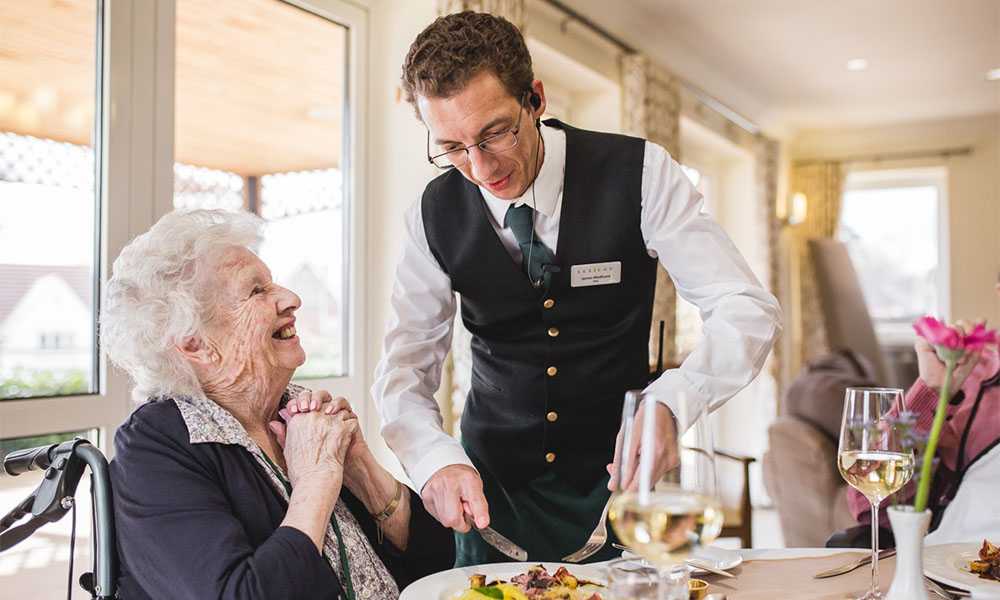 Young waiter serving food to happy, seated older lady
