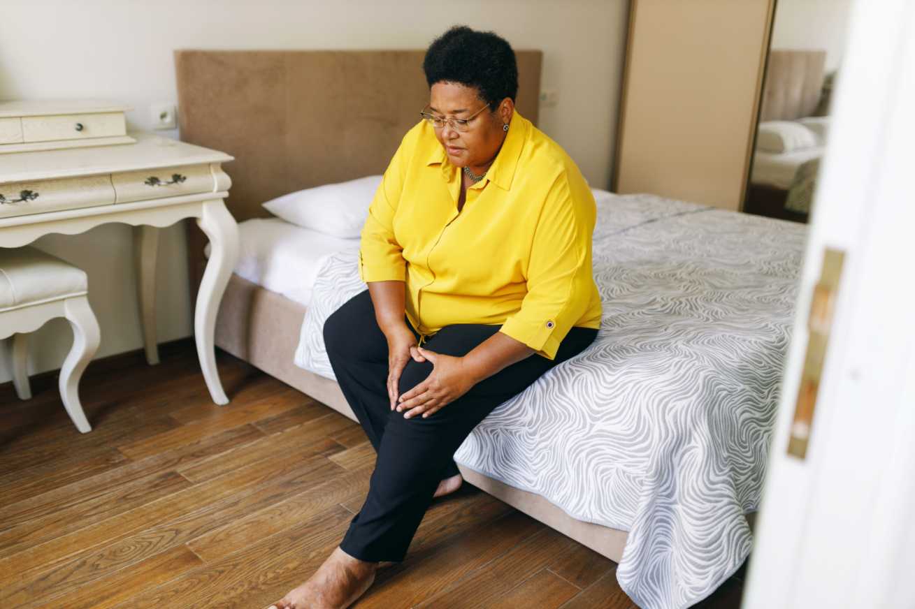 Older woman in pain trying to get off her bed, the woman wears a bright yellow shirt and black trousers and holds her knee in pain